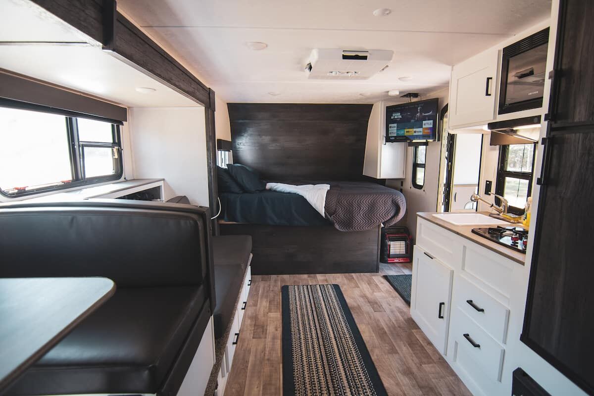 Inside the Houston Camper: a bed with a wood headboard, a dark sofa, white cabinets and a kitchenette