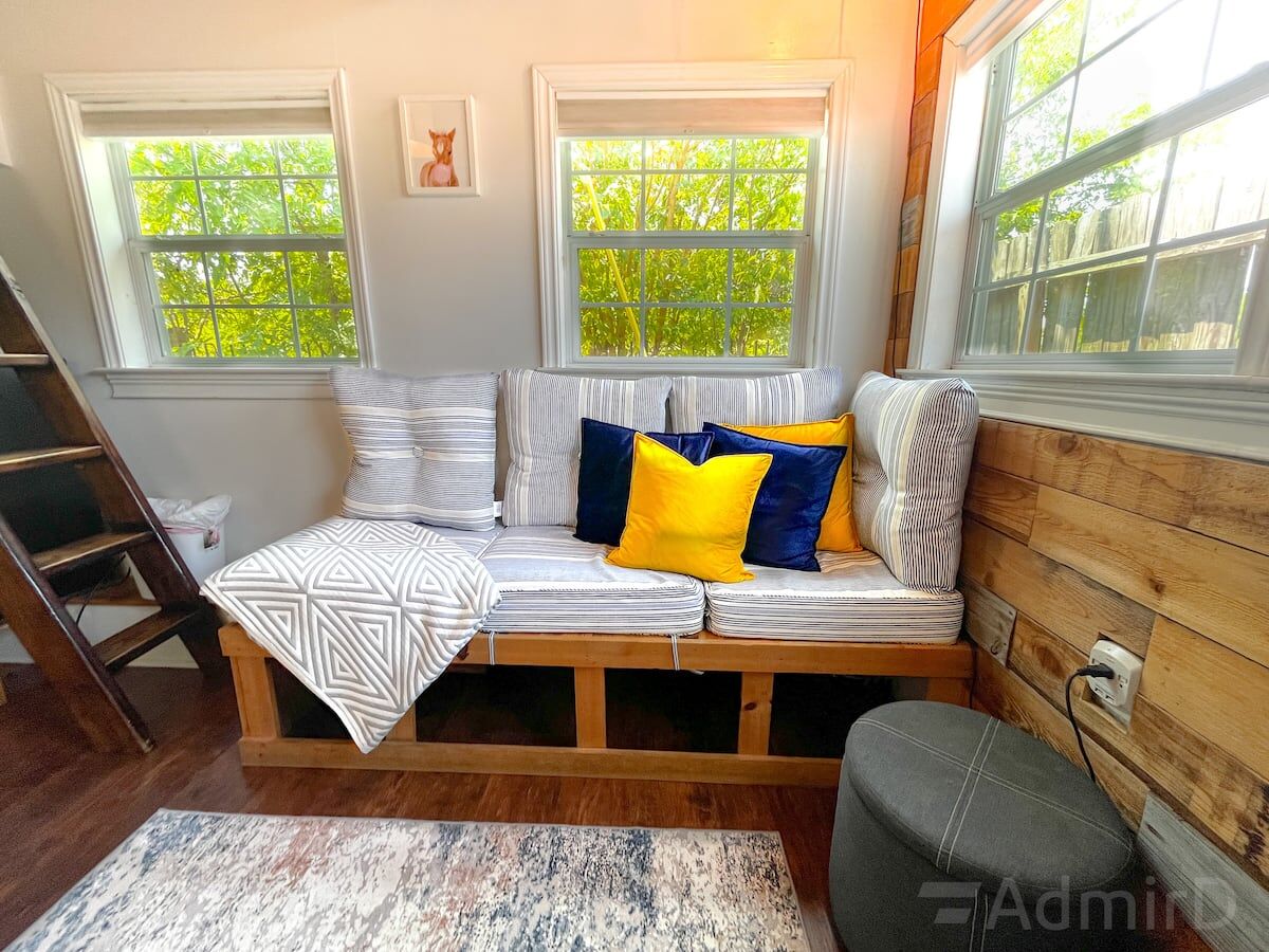Sunlit living area in the Houston Tiny House with a built-in bench sofa, yellow and navy pillows and wood-paneled walls